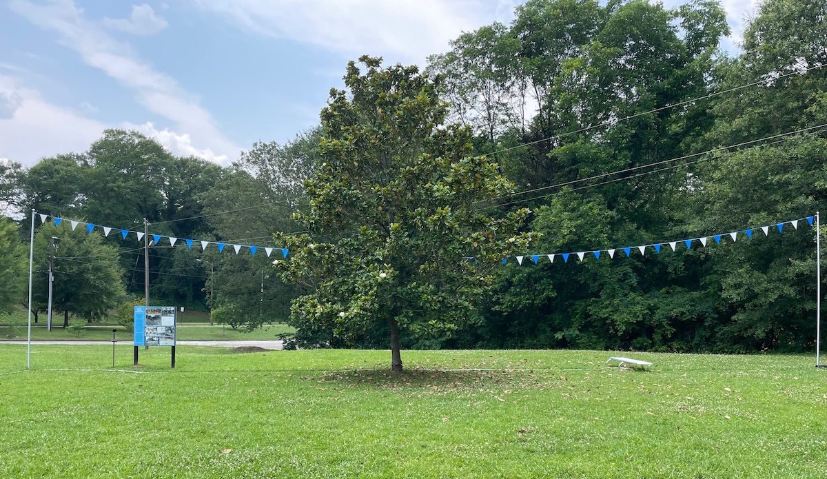 Photo shows a grassy field that once was the Spring Street pool. The pool is outlined in pavers, and the diving board has been returned to its original spot. Pool flags also fly across where the pool once was.