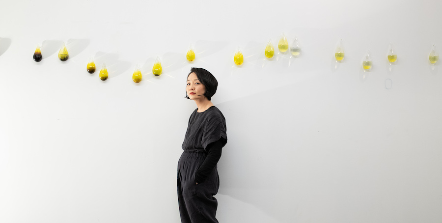 Artist Gyun Hur in black standing in front of handblown tear-shaped vessels in shades of yellow and black hanging on the wall in her studio