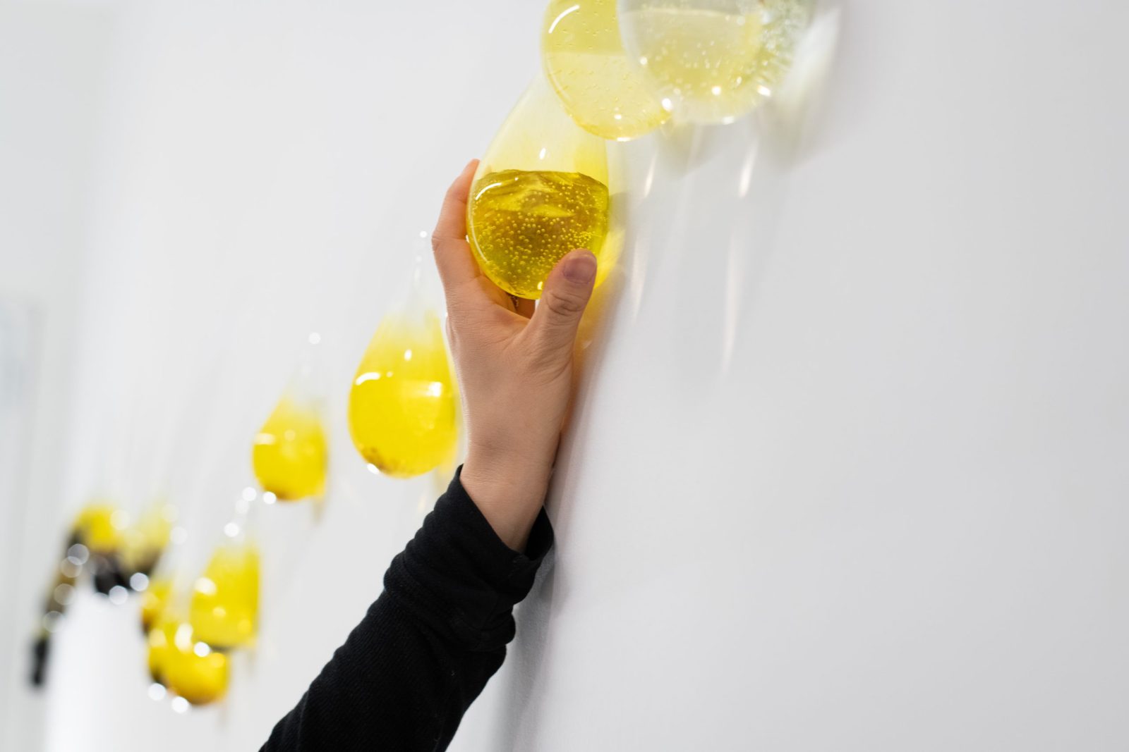 Photo shows Gyun's hand hanging one of the handblown tear-shaped vessels on a white wall.