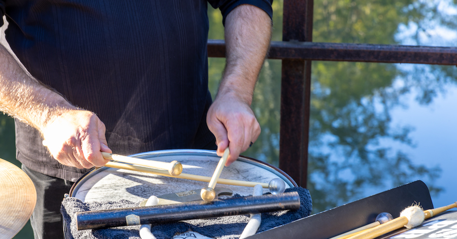 Hands of a musician seen playing a drum with water in the background. Photo by John Ramspott